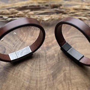 May include: Two brown leather bracelets with rectangular metal clasps. The bracelets are circular and sit on a wooden surface. The leather is a rich, dark brown colour, and the metal clasps are silver and black.
