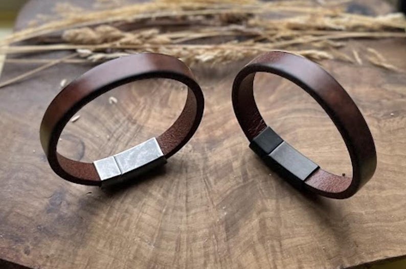 May include: Two brown leather bracelets with rectangular metal clasps. The bracelets are circular and sit on a wooden surface. The leather is a rich, dark brown colour, and the metal clasps are silver and black.