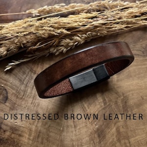 Puede incluir: Una pulsera de cuero marrón desgastado con un cierre plateado. La pulsera se muestra sobre una superficie de madera con plantas secas en el fondo. El texto en la imagen dice "NEW DISTRESSED BROWN LEATHER."