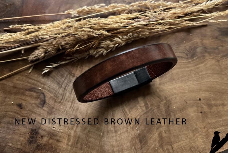 May include: A brown leather bracelet with a distressed finish and a silver clasp. The bracelet is shown on a wooden surface, with dried plants in the background. The image includes the text "NEW DISTRESSED BROWN LEATHER."
