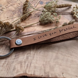 Può includere: Un portachiavi in ​​cuoio marrone con un anello per chiavi in ​​argento. Il portachiavi ha le parole "Ti voglio bene Nonno" scritte in inchiostro nero.