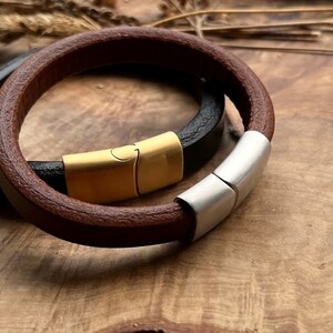 Puede incluir: Dos pulseras de cuero con cierres magnéticos. Una pulsera es de cuero marrón con un cierre plateado, y la otra es de cuero negro con un cierre dorado.