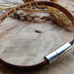 Puede incluir: Un brazalete de cuero marrón con un cierre plateado. El brazalete está grabado con las palabras "You are always on my mind".
