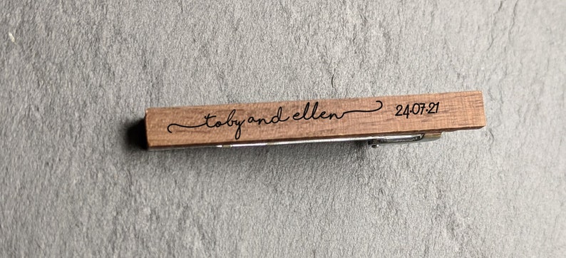 May include: A wooden tie clip with the text "Toby and Ellen" and the date "24.07.21" engraved on it.