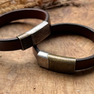 May include: Two brown leather bracelets with silver and brass clasps. The bracelets are on a wooden surface.