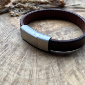 May include: A brown leather bracelet with a silver clasp. The bracelet is on a wooden surface.