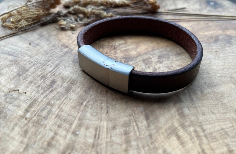 May include: A brown leather bracelet with a silver clasp. The bracelet is on a wooden surface.