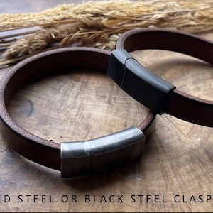 May include: Two brown leather bracelets with brushed steel or black steel clasps. The bracelets are circular and lie on a wooden surface, with dried plants in the background. The clasps are rectangular and secure the bracelets.