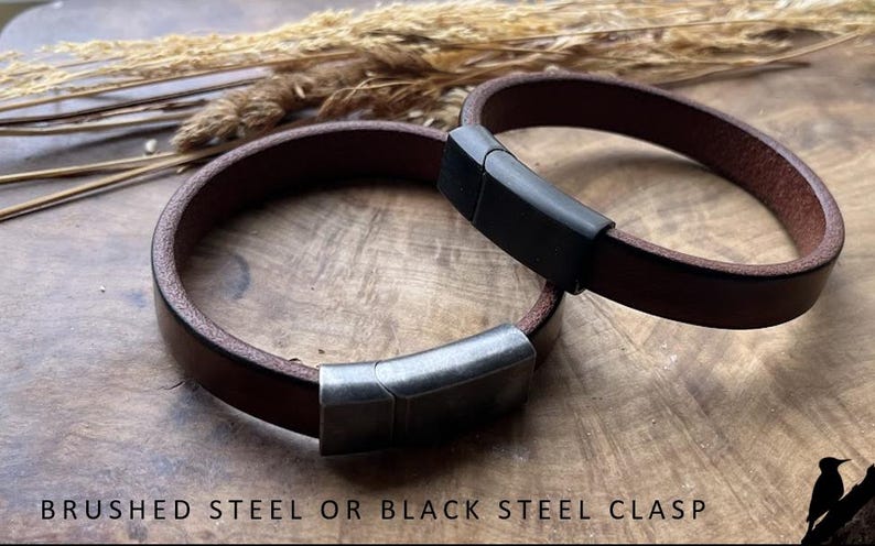 May include: Two brown leather bracelets with brushed steel or black steel clasps. The bracelets are circular and lie on a wooden surface, with dried plants in the background. The clasps are rectangular and secure the bracelets.