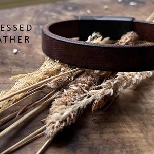 May include: A dark brown leather bracelet with a black clasp, displayed on a wooden surface. The text "NEW DISTRESSED BROWN LEATHER" is visible. The bracelet is surrounded by dried plants, creating a rustic aesthetic.