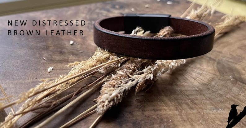 May include: A dark brown leather bracelet with a black clasp, displayed on a wooden surface. The text "NEW DISTRESSED BROWN LEATHER" is visible. The bracelet is surrounded by dried plants, creating a rustic aesthetic.