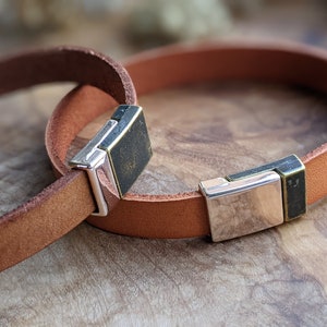 Peut inclure: Deux bracelets en cuir marron avec des fermoirs en métal argenté et noir. Les bracelets sont empilés l'un sur l'autre.