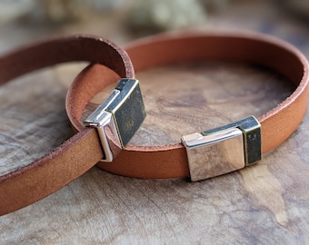Könnte beinhalten: Zwei braune Lederarmbänder mit silbernen und schwarzen Metallschließen. Die Armbänder liegen übereinander.