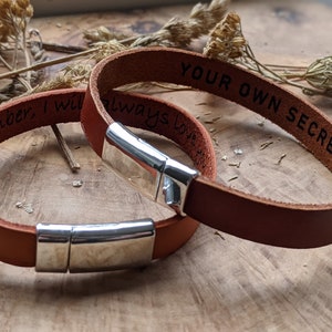 Puede incluir: Dos pulseras de cuero marrón con cierres plateados. Una pulsera está grabada con "Remember, I will always love you" y la otra con "Your own secret mess".