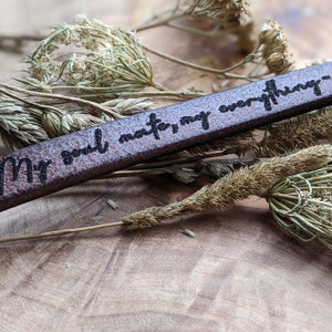 May include: A brown leather bracelet with the text "My soul mate, my everything - love" engraved on it. The bracelet is lying on a bed of dried flowers.