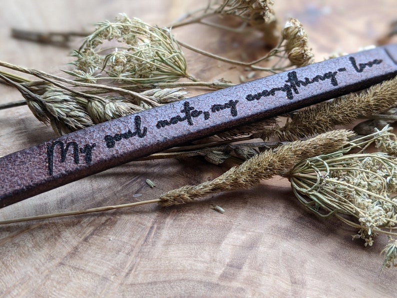 May include: A brown leather bracelet with the text "My soul mate, my everything - love" engraved on it. The bracelet is lying on a bed of dried flowers.