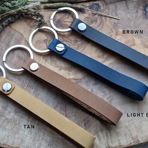 Könnte beinhalten: Vier Leder-Schlüsselanhänger in verschiedenen Farben: Braun, Hellbraun, Marineblau und Braun. Jeder Schlüsselanhänger hat einen silbernen Schlüsselring und eine silberne Metallschaufel.