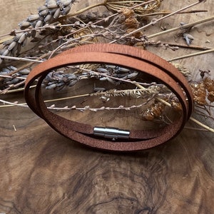 Peut inclure: Un bracelet en cuir marron avec une fermeture magnétique argentée. Le bracelet est posé sur une surface en bois avec des fleurs séchées.