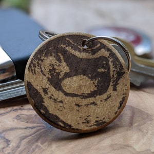 Op de afbeelding: Een ronde houten sleutelhanger met een donkerbruin ontwerp. De sleutelhanger is bevestigd aan een zilveren sleutelring. Het ontwerp is een abstract patroon. Andere sleutels en een zwart object staan op de achtergrond.