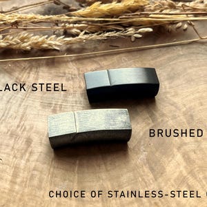 May include: Two stainless steel clasps, one black and one brushed steel, are displayed on a wooden surface. The text "BLACK STEEL" and "BRUSHED STEEL" are above each clasp. The text "CHOICE OF STAINLESS-STEEL CLASPS" is below.