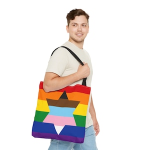 Op de afbeelding: Een vierkante draagtas met een horizontaal regenboogstrepenpatroon. In het midden van de tas staat een zespuntige ster gevuld met horizontale strepen in bruin, zwart, lichtblauw, roze en wit. De tas heeft twee zwarte schouderbanden.