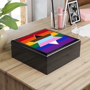 May include: A square black wooden keepsake box featuring a flat lid with a printed design. The design displays a horizontal rainbow striped background overlaid with a large, centered six-pointed star colored in light blue, pink, and white.