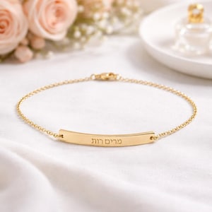 Può includere: Un braccialetto dorato con una catena delicata e una barra curva. La barra presenta un testo inciso. Il braccialetto è esposto su una superficie bianca con morbidi accenti floreali sullo sfondo. È visibile anche un piccolo flacone di profumo.