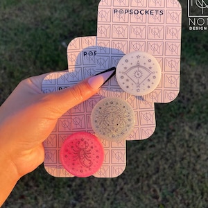Könnte beinhalten: Ein Set aus drei PopSockets mit Tierkreiszeichen-Designs. Das Set enthält einen pinkfarbenen Skorpion, einen glitzernden silbernen Krebs und einen weißen Schützen. Die PopSockets werden auf einer Karte mit dem Text "POPSOCKETS" und "NONY DESIGN + CO" präsentiert.