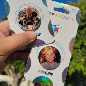 Puede incluir: Dos PopSockets para teléfono con detalles holográficos. Uno presenta una foto de una mujer con dos perros, el otro un retrato de una mujer. El embalaje incluye el texto "POPSOCKETS" y "POPGRIP".
