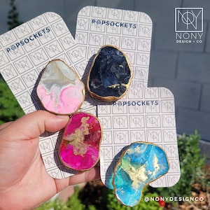 Könnte beinhalten: Vier PopSockets mit einzigartigen Designs. Einer hat rosa und weiße Wirbel, ein anderer ist dunkelblau mit goldenen Akzenten, ein dritter ist pink mit Gold und der letzte ist blau und grün mit Gold. Jeder hat einen goldfarbenen Rand und wird auf einer Markenkarte präsentiert.
