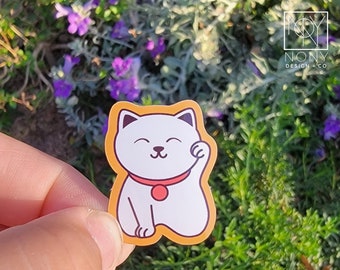 Könnte beinhalten: Ein weißer Maneki-neko-Aufkleber mit rotem Kragen und erhobener Pfote, umrandet von einem orangefarbenen Rand. Der Aufkleber zeigt ein lächelndes Katzendesign. Der Aufkleber wird in einer Hand gehalten, mit einem Hintergrund aus grünem Laub und lila Blumen.
