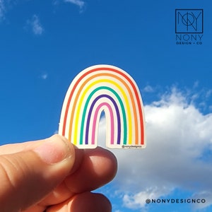 May include: A colorful rainbow sticker with a white background and a curved shape. The rainbow features stripes of red, orange, yellow, green, blue, and purple. The sticker is held against a blue sky background. The text "@nonydesignco" is visible.