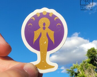 Puede incluir: Una pegatina con la silueta dorada de una figura con los brazos extendidos, adornada con estrellas y una luna creciente, sobre un fondo morado con fases lunares. La pegatina tiene un borde blanco y el texto "NONY DESIGN + CO".