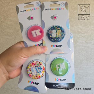 Op de afbeelding: Vier PopSockets telefoonhouders in roze, blauw en groen, elk met glitter en initialen. De verpakking leest "POPSOCKETS" en "POPGRIP". De merknaam "NONY DESIGN + CO" is zichtbaar.