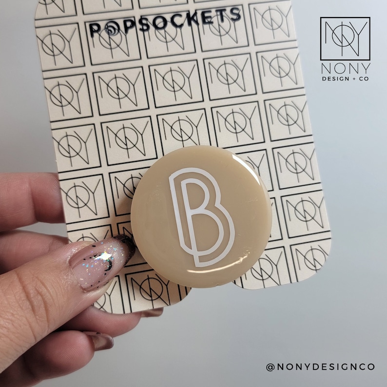 May include: A beige popsocket with a white letter B in the center. The popsocket is on a white background with a repeating pattern of black squares with a white line inside. Custom Initial PopSocket™: Personalized Phone Grip, Resin Art