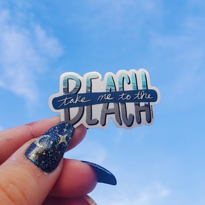 May include: A white sticker with the words "BEACH" and "take me to the BEACH" in blue and gray lettering. The word "BEACH" has a watercolor ocean design. The sticker is held against a bright blue sky.