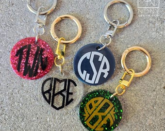 May include: Keychains with custom initials in various colors and shapes. The keychains include a pink glitter circle with black initials, a clear heart with black initials, a navy blue circle with white initials, and a green glitter circle with gold initials.