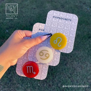 May include: Three PopSockets with zodiac symbols: a red Scorpio, a yellow Leo, and a white Cancer. Each PopSocket is attached to a patterned card with the brand name. The background is a grassy area.