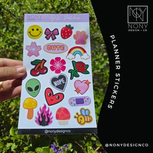 May include: A white sticker sheet titled NONY DESIGN + CO Y2K Patches featuring various colorful designs including a yellow smiley face, pink bow, red strawberry, paw print, cherry, rainbow, alien head, heart with NOPE text, rose, and bee.