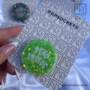 Könnte beinhalten: Ein grüner Glitzer-PopSocket mit den Worten "DEAD INSIDE" in weißer Schrift. Der PopSocket befindet sich auf einer weißen Karte mit einem schwarzen geometrischen Muster und den Worten "POPSOCKETS". Der Markenname "NONY DESIGN + CO" ist ebenfalls sichtbar.