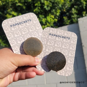 Könnte beinhalten: Zwei runde PopSockets mit strukturierten Oberflächen, einer hellbraun und einer dunkelgrau, werden auf Karten mit dem Wort "POPSOCKETS" und einem sich wiederholenden Muster präsentiert. Der Markenname "NONY DESIGN CO" ist rechts oben sichtbar.