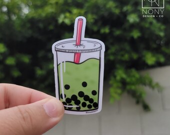 May include: A sticker featuring a cartoon illustration of a green bubble tea drink with black tapioca pearls. The drink is in a clear plastic cup with a white lid and a red straw. The sticker has the text "NONY DESIGN + CO" and "@NONYDESIGNCO".