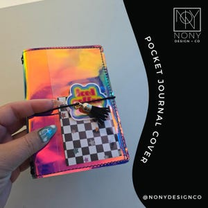 May include: A rectangular pocket journal cover with a reflective, iridescent finish. The cover features a black and white checkered insert with small star accents, a sticker reading iced coffee, and a black elastic band with a black tassel charm.