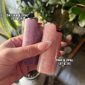 以下が含まれることがあります： ラベンダーとピンクの2つのカスタムライター。グリッターとオパールアクセント付き。画像には「DATURA & OPAL」と「PINK & OPAL」と書かれています。ライターは、赤い点火ボタンが付いた標準的な長方形の形状をしています。