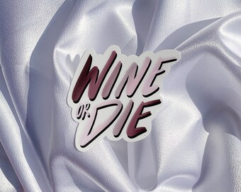 May include: A sticker with the phrase "WINE OR DIE" in a handwritten style. The text is in shades of pink and burgundy, outlined in black, and set against a white background. The sticker is placed on a white, silky fabric.