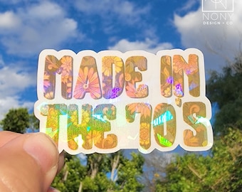Può includere: Un adesivo olografico con la scritta "MADE IN THE 70'S" in un font retrò. Le lettere sono riempite con un motivo floreale nei toni dell'arancione, del rosa e del marrone. L'adesivo è tenuto contro uno sfondo di cielo blu e fogliame verde.