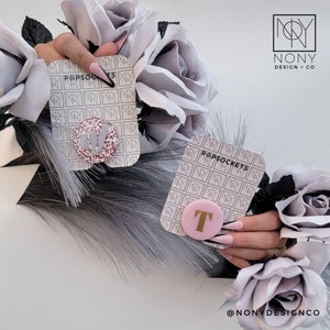 Op de afbeelding: Twee PopSockets met initialen, &eacute;&eacute;n glitterroze met een letter en de andere roze met een letter. De PopSockets worden getoond met grijze rozen en veren. De merknaam "NONY DESIGN + CO" is zichtbaar.