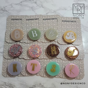 Op de afbeelding: Een verzameling PopSockets met verschillende initialen. De PopSockets zijn rond en hebben verschillende kleuren, waaronder beige, groen, roze en bruin. Sommige hebben glitteraccenten. De letters zijn goud of wit. De merknaam "NONY DESIGN + CO" is zichtbaar.