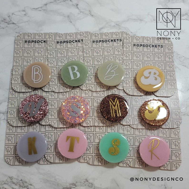 Op de afbeelding: Een verzameling PopSockets met verschillende initialen. De PopSockets zijn rond en hebben verschillende kleuren, waaronder beige, groen, roze en bruin. Sommige hebben glitteraccenten. De letters zijn goud of wit. De merknaam "NONY DESIGN + CO" is zichtbaar.