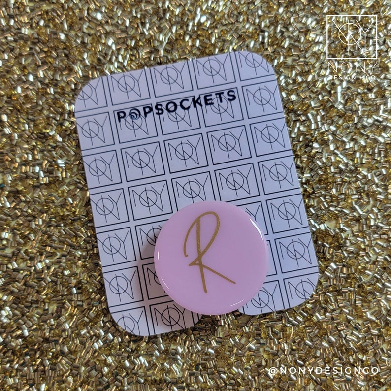 Op de afbeelding: Een roze PopSocket met een gouden letter "R" op een zwart-wit patroonkaart. De kaart leest "POPSOCKETS" in zwarte tekst. De achtergrond is een gouden glitteroppervlak.
